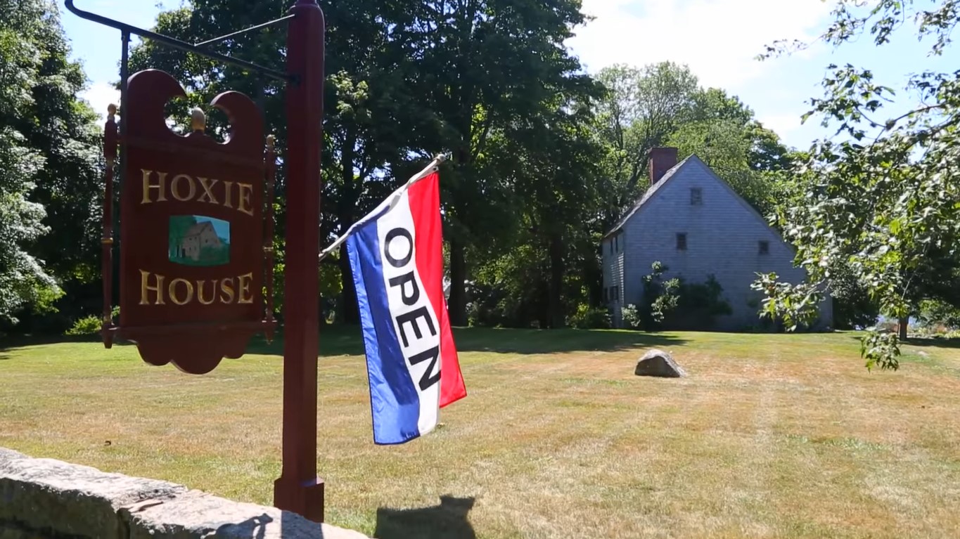A dark brown ‘Hoxie House’ sign hanging from a post in a yard and a flag that says ‘Open’