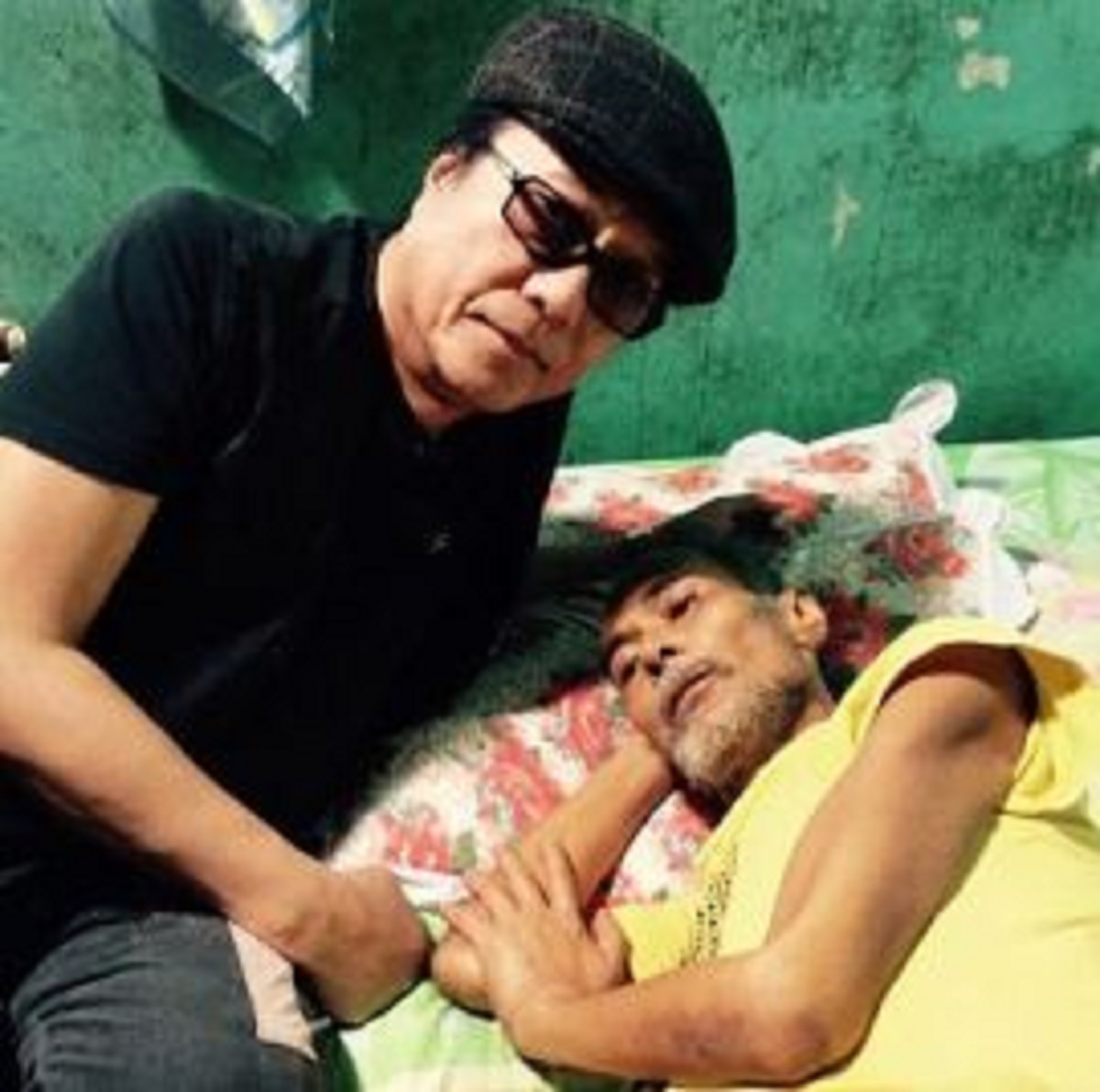 Freddie Aguilar in black T-shirt and beret sitting beside Roel Cortez in yellow T-shirt and lying on his bed